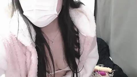 Media: Video of an Asian woman with long black hair, wearing a white surgical mask, pink fur-trimmed jacket, and a revealing white lace bra.