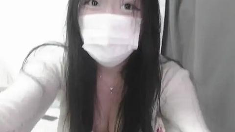 Media: Video of an East Asian woman with long black hair, wearing a white face mask, revealing a cleavage and necklace, indoors with blurred background.