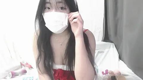 Media: Video of an East Asian woman with long black hair, wearing a red dress, white mask, and a cross necklace, adjusting her hair, surrounded by toys and a bed in a dimly lit room.