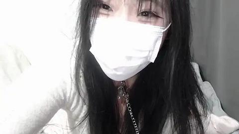 Media: A close-up video of an Asian woman with long black hair, wearing a white surgical mask and a dark top. She has a slender build and a neutral facial expression.