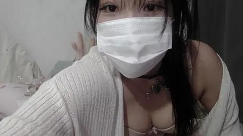 Media: Video of an Asian woman with long black hair, wearing a white face mask, a beige bra, and a white knitted cardigan. The background is a dimly lit room with white walls and a bed.