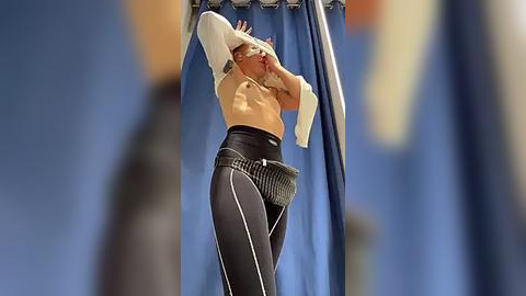 Media: Video of a tall, muscular woman with light skin, wearing a white sports bra and black leggings, stretching in a gym with blue curtains in the background.