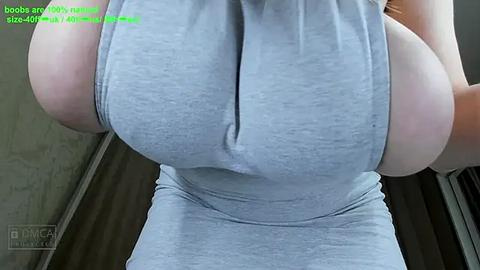 Media: A close-up video of a woman's large breasts, partially obscured by a tight, light gray tank top, with a green text overlay and a watermark in the bottom left corner.