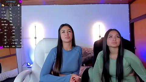 Media: Video of two young women with long dark hair, dressed in blue and green tops, sitting on white chairs in a cozy, dimly-lit room with orange walls and wooden ceiling.