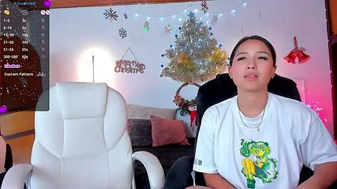 Media: Video of a young woman with a tan complexion, dark hair in a ponytail, wearing a white graphic T-shirt, sitting in a white office chair, in a festive room with a Christmas tree, decorations, and a \"Merry Christmas\" sign.