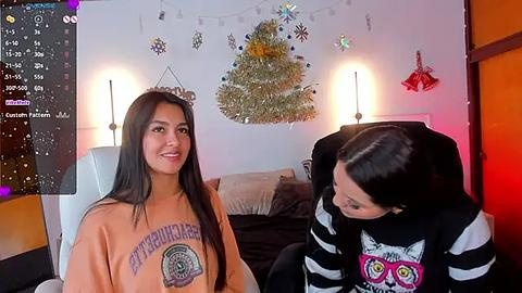 Media: Video of a young woman with long black hair, wearing a \"Burlington\" sweatshirt, sitting on a white chair in a cozy room with a Christmas tree, string lights, and a black cat-themed sweater.