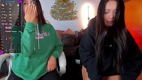 Media: Video of two young women in a cozy living room, one in a green hoodie, the other in black, both laughing. Background features a TV screen with streaming content and a Christmas tree.