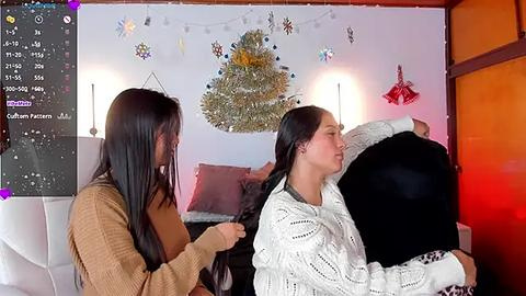 Media: Video of two women in a cozy bedroom, one with long hair being braided, the other wrapped in a white knit sweater. Christmas decorations adorn the white wall.