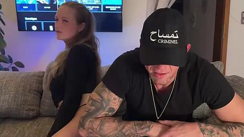 Media: Video of a tattooed man in a black cap, leaning on a couch, watching TV; a blonde woman with long hair sits beside him.