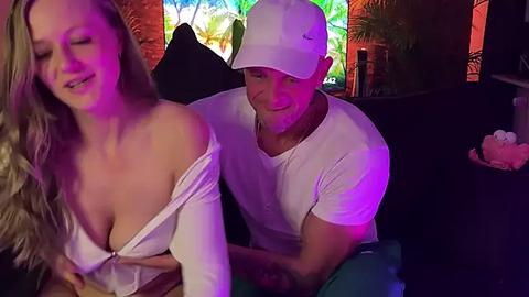 Media: Video of a blonde woman with large breasts in a white off-shoulder top and a muscular man in a white cap and shirt, both smiling at each other in a dimly-lit bar.