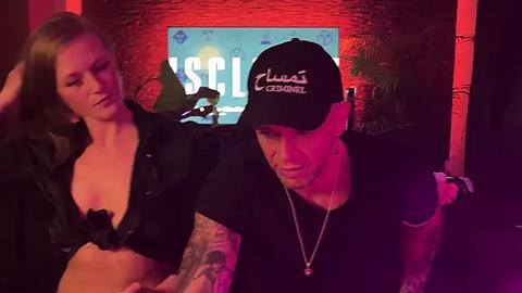 Media: Video of a tattooed, muscular man in a black cap and shirt, leaning over a blonde woman in a revealing black top, in a dimly lit, red-lit room with a \"USC\" sign and plant in the background.