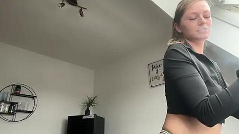 Media: A video of a woman with fair skin and light brown hair, wearing a black crop top, standing in a modern, minimalist room with a white wall and a circular mirror.