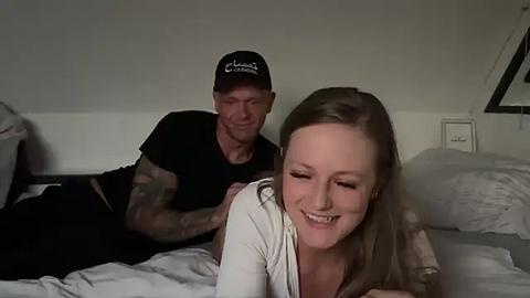 Media: A video captures a tattooed man in a black cap and shirt, smiling, leaning over a blonde woman with a white shirt, laughing. They are in a dimly lit bedroom with white walls and a bed.