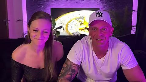 Media: Video of a smiling, tattooed man in a white t-shirt and Yankees cap, seated next to a blonde woman in a black glittery top, in a dimly-lit room with a TV displaying a retro clock.