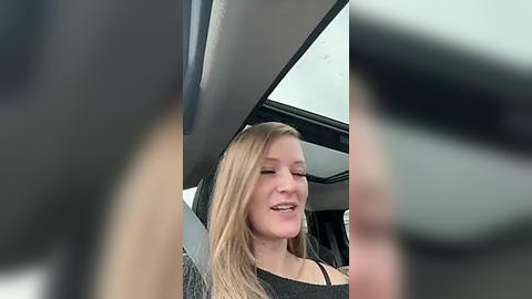 Media: Video of a smiling young blonde woman with straight hair, wearing a black top, sitting in a car, viewed through a blurred, black-framed window.