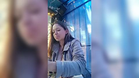 Media: Video of a young woman with long brown hair, wearing a light jacket, seated inside a glass-walled, blue-tinted booth at a restaurant. Background features blurred figures and warm yellow lights.