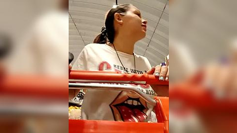 Media: Video of a young woman with dark hair in a ponytail, wearing a white t-shirt with red text, holding a red cup, standing in a modern, arched-ceiling room.