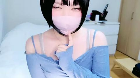 Media: A video of a slender, pale-skinned Asian woman with short black hair and bangs, wearing a light blue off-shoulder sweater and a pink mask, holding her finger to her lips in a bedroom with white furniture and a bed.