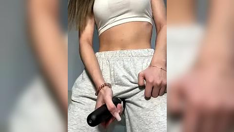 Media: Video of a light-skinned woman in a white sports bra and gray sweatpants, holding a black mascara wand, with blurred figures in the background.