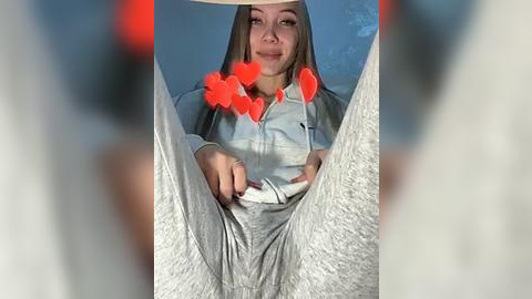 Media: Video of a young woman with light skin, long brown hair, and a gray hoodie, legs spread, heart emojis covering her face, blurred background.