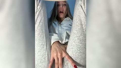 Media: Video of a young woman with long blonde hair, wearing a light gray hoodie, legs spread, reaching down, revealing her vagina. Background is a blurred blue curtain.