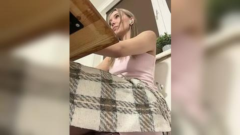 Media: Video of a blonde woman in a pink tank top and checkered skirt, leaning over a wooden table, with a blurred background.