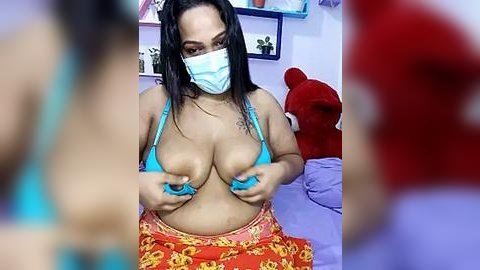 Media: Video of a Latina woman with medium skin tone, long black hair, wearing a blue mask, turquoise bikini top, and red skirt with yellow flowers, holding her large breasts. Background includes a red teddy bear and blue shelves with plants.