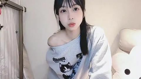 Media: Video of a young Asian woman with long black hair, fair skin, and bangs, wearing a light gray off-shoulder sweatshirt with black cat print, sitting on a white leather couch, in a softly lit room.