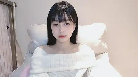 Media: A video of an Asian woman with long black hair and fair skin, wearing a white sweater, lying on a white cushioned chair in a minimalist room.