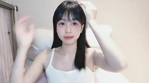 Media: Video of an Asian woman with long black hair, wearing a white tank top, sitting on a bed, holding her hair, and smiling.