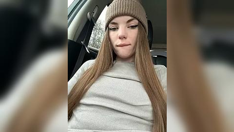 Media: Video of a young Caucasian woman with long, straight, light brown hair, wearing a beige knit beanie and a light gray sweater, seated in a car with blurred exterior background.