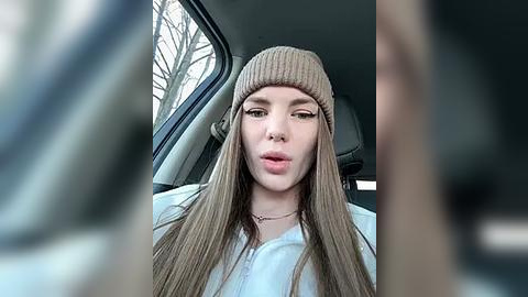 Media: Video of a young woman with long, straight, brown hair, wearing a beige knitted beanie and a white sweater, seated in a car with blurry, leafless trees outside the window.