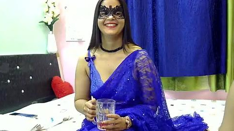 Media: Video of a South Asian woman with medium skin tone, long black hair, and a black mask, wearing a blue sari and black choker, holding a glass, smiling in a bedroom with blue curtains and white bed.