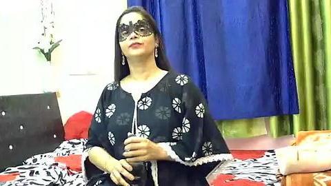 Media: Video of a woman with long black hair, wearing a black mask and black floral-patterned blouse, sitting on a bed with red and white sheets, blue curtains, and green drapes in the background.