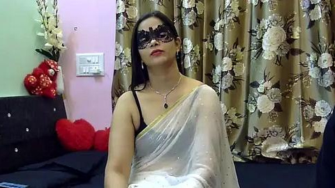 Media: Video of a woman in a white saree, black lace mask, and black bra, sitting on a black sofa with red pillows and floral curtains in a room.