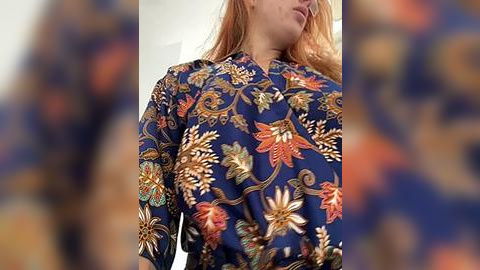 Media: Video of a woman with long, light brown hair, wearing a blue blouse adorned with intricate, gold floral embroidery. The background is blurred, emphasizing the colorful details of the blouse.