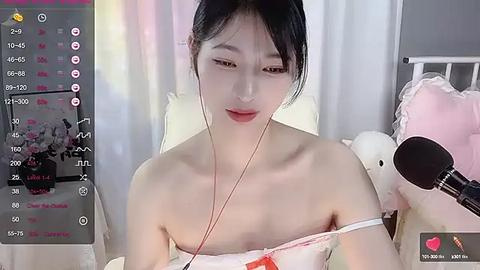 Media: A video of a young East Asian woman with fair skin and dark hair, wearing an off-shoulder white dress, listening to music with headphones, in a cozy bedroom setting.