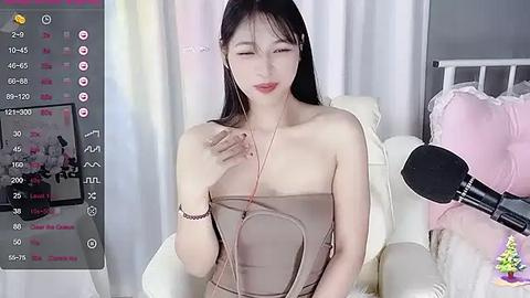 Media: A video of an East Asian woman with long black hair, wearing a strapless beige dress, sitting in a white chair, smiling, with a pink microphone nearby.