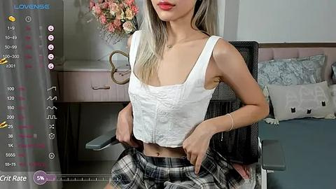 Media: Video of a slender, light-skinned woman with long, blonde hair, wearing a white crop top and plaid skirt, sitting in a modern bedroom.