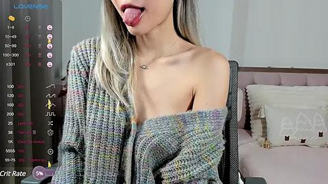 Media: Video of a young, light-skinned woman with long blonde hair, wearing a loose, off-shoulder sweater. She sticks out her tongue, posing seductively in a bedroom with a pink bed and white pillows.