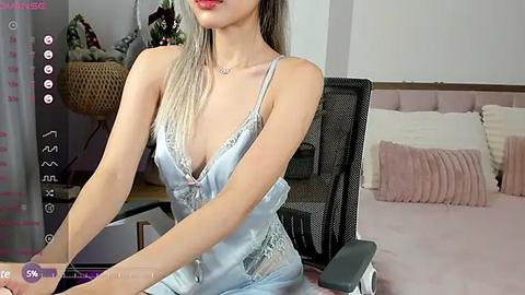 Media: A video of an East Asian woman with long, straight, platinum blonde hair, wearing a silky, light blue camisole with lace trim, sitting in a modern black office chair in a softly lit bedroom with a neatly made bed.