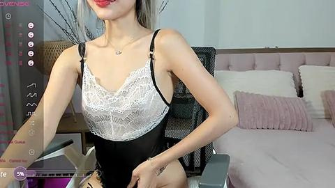 Media: Video of an Asian woman with fair skin, straight blonde hair, and red lipstick, wearing a black lace bra and panties, sitting in a modern bedroom with a bed, pink pillows, and a black mesh chair.