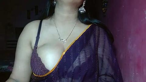 Media: A video of a woman with light skin, wearing a purple saree with a gold border, and a sheer purple bra, revealing ample cleavage, standing in a dimly lit room with pink walls and indistinct furniture in the background.