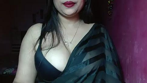 Media: Video of a light-skinned woman with long black hair, wearing a black sheer top that reveals a black bra, red lipstick, and a gold necklace. Background shows a purple wall and indistinct objects.