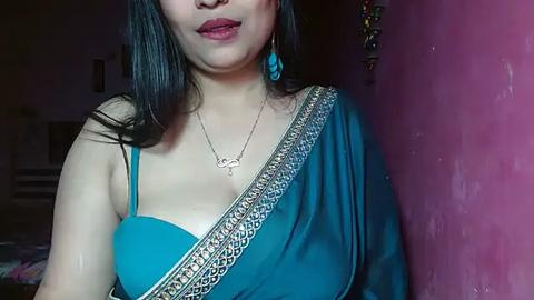 Media: Video of a South Asian woman with medium skin tone, wearing a teal sari with intricate gold border, blue earrings, and a necklace. Background shows a pink wall and a dimly lit room.