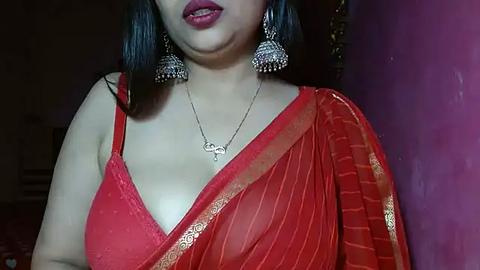 Media: Video of a South Asian woman with medium skin tone, dark hair, and bold red lipstick, wearing a red saree with gold embroidery, and silver chandelier earrings, indoors with a purple wall.