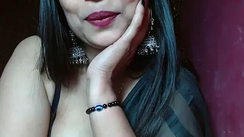 Media: Video of a woman with long black hair, wearing a dark blouse, large silver earrings, and a black beaded bracelet. She has light skin, dark lipstick, and rests her chin on her hand.