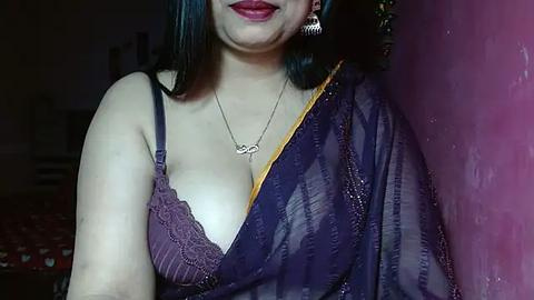 Media: Video of a woman with medium skin tone, wearing a purple saree with a lace bralette visible, adorned with a silver necklace and earrings, standing against a pink wall in a dimly lit room.