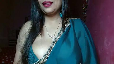 Media: Video of a South Asian woman with long black hair, fair skin, and a medium build, wearing a teal sari with gold border. She has red lipstick, a gold necklace, and turquoise earrings. Background is dimly lit, with a red wall and a blurred room.