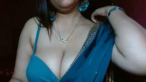 Media: Video of a light-skinned woman with medium-length dark hair, wearing a low-cut blue saree with gold detailing, blue earrings, and a silver necklace, posing in a dimly lit room.